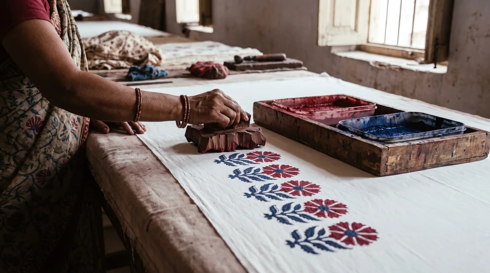 Artisan hand-stamping a fine red floral buti onto white cotton with a carved wooden block on a Sanganer printing table